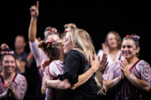 KJ Kindler hugs an Oklahoma gymnast while the team cheers in the background.