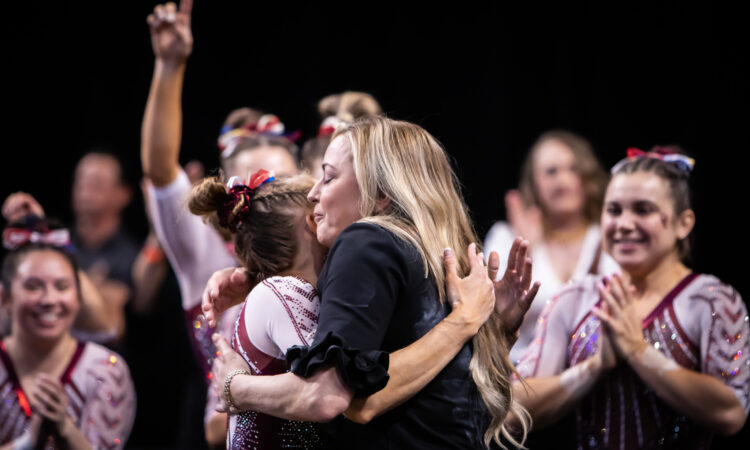 KJ Kindler hugs an Oklahoma gymnast while the team cheers in the background.