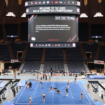 a shot from high up in the stands of the whole arena floor at the Big 12 championships during warmups. Gymnasts are running around the floor and stretching, and the jumbotron is at the top of the shot.