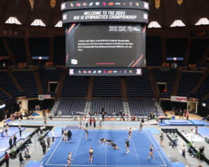 a shot from high up in the stands of the whole arena floor at the Big 12 championships during warmups. Gymnasts are running around the floor and stretching, and the jumbotron is at the top of the shot.