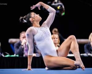 Kayle DiCello sits on one leg on the floor with her other lag crossed over. She leans on on hand and is looking up towards her other outstretch palm as she performs her floor routine