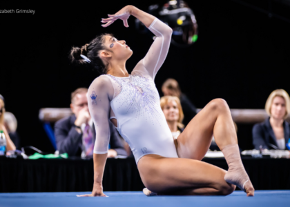 Kayle DiCello sits on one leg on the floor with her other lag crossed over. She leans on on hand and is looking up towards her other outstretch palm as she performs her floor routine