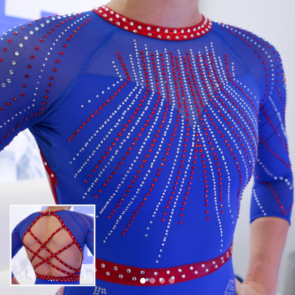 A gymnast wearing a blue leotard with silver and red rhinestones radiating from the neckline and down the sleeves. The back features a mesh design with crisscrossing straps in a pattern for an elegant and sleek look.
