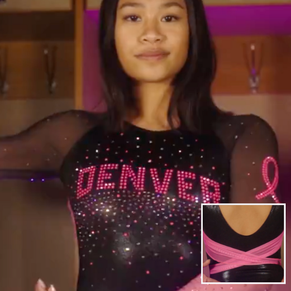 A gymnast from the University of Denver poses in a black leotard with bright pink details. The front has "DENVER" spelled out in pink rhinestones, surrounded by shimmering stones, with a breast cancer ribbon on the sleeve. The back has a crisscross pink strap detail against a deep V-neckline. The overall theme is breast cancer awareness, with elegant, sparkling embellishments.