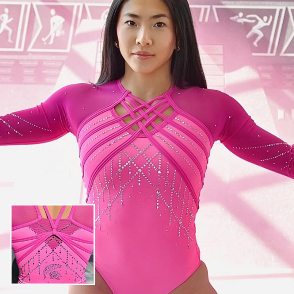 A Michigan State gymnast models the team’s new pink leotard. The design features dark magenta at the shoulders and bright pink on the body, accented with sparkling rhinestones in a geometric pattern. The neckline showcases a crisscross strap detail, and the sleeves are adorned with scattered rhinestones. An inset image highlights the back, which has layered strap details and additional rhinestone embellishments forming the Spartan helmet logo near the lower back.