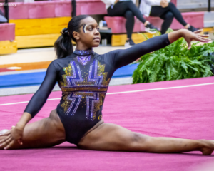 Gymnast performing a floor routine in a black leotard with golden accents, extending one arm gracefully while seated on a maroon mat, surrounded by plants and bleachers in the background.