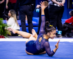 A gymnast performing a graceful floor routine, showcasing flexibility and precision, wearing a navy leotard with sheer sleeves and yellow accents. Her hair is styled in a bun with a yellow ribbon, and the routine exudes elegance and confidence. Photo credit: Tara Graeve.