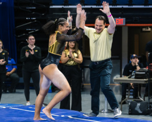 A gymnast and coach celebrate with a high-five during a meet, showcasing energy and camaraderie. The gymnast is wearing a gold and black leotard, while the coach is dressed in a matching team polo, both surrounded by cheering teammates and staff in the background. Photo credit: Hope Maier.