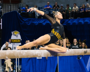 Lauren Keener of Towson performs a graceful split on the balance beam, showcasing her precision and poise during a competition. The black and gold checkered design of her leotard highlights the Towson team spirit. (Photo by Hope Maier)