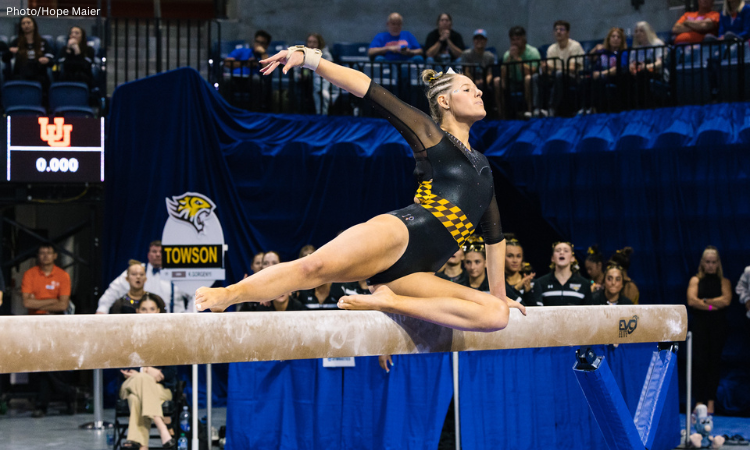 Lauren Keener of Towson performs a graceful split on the balance beam, showcasing her precision and poise during a competition. The black and gold checkered design of her leotard highlights the Towson team spirit. (Photo by Hope Maier)