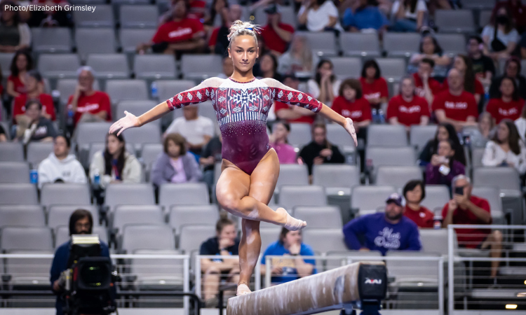 Female gymnast wearing a red and white leotard performs a balance beam routine in an arena filled with spectators, showcasing precision and focus. Photo credit: Elizabeth Grimsley.