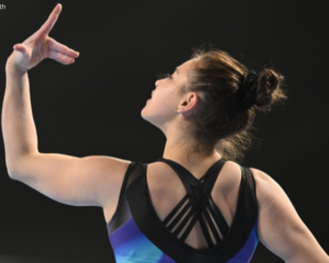 An artistic gymnastics athlete in mid-pose, showcasing grace and elegance, with hair in a bun, wearing a colorful leotard with black strap detailing on the back, captured against a dark background. Photo credit: Lloyd Smith.