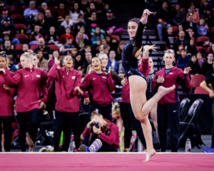 A gymnast with long, dark hair pulled back into a ponytail is captured mid-performance on a red floor. She is wearing a black leotard and has one leg extended behind her. Behind her, a group of at least eight teammates are watching. They are wearing matching maroon jackets with a white logo on the chest. Their expressions range from anticipation to encouragement. One person in the foreground is kneeling and appears to be taking a photograph.