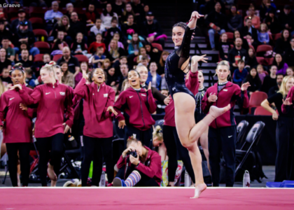 A gymnast with long, dark hair pulled back into a ponytail is captured mid-performance on a red floor. She is wearing a black leotard and has one leg extended behind her. Behind her, a group of at least eight teammates are watching. They are wearing matching maroon jackets with a white logo on the chest. Their expressions range from anticipation to encouragement. One person in the foreground is kneeling and appears to be taking a photograph.
