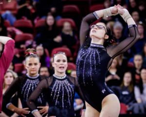 A woman gymnast with glasses, wearing a black leotard with sparkly embellishments, performing on floor during a gymnastics competition.