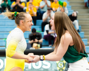 A female gymnast with blonde hair in a ponytail, wearing a white and yellow leotard, smiles and clasps hands with another smiling woman with long brown hair, wearing a green tank top and white pants. They are both looking at each other, appearing excited. In the background, out of focus, are spectators in bleachers.