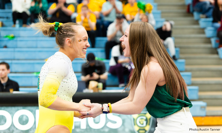 A female gymnast with blonde hair in a ponytail, wearing a white and yellow leotard, smiles and clasps hands with another smiling woman with long brown hair, wearing a green tank top and white pants. They are both looking at each other, appearing excited. In the background, out of focus, are spectators in bleachers.