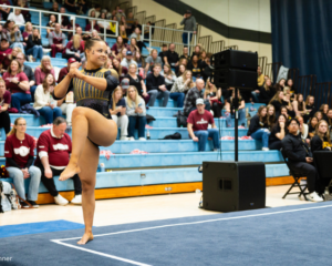 Rhode Island gymnast Olivia Keyes performs on floor exercise during a meet, smiling with her arms crossed and one leg raised mid-dance. She wears a navy and gold leotard, with a full crowd and judges visible in the background.