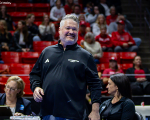Southern Utah gymnastics coach Scotty Bauman smiles as he stands near the competition floor, wearing a black Southern Utah pullover. He is surrounded by judges and staff, with a crowd of spectators seated in red bleachers in the background.
