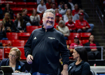 Southern Utah gymnastics coach Scotty Bauman smiles as he stands near the competition floor, wearing a black Southern Utah pullover. He is surrounded by judges and staff, with a crowd of spectators seated in red bleachers in the background.