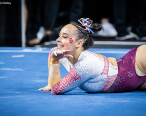 Faith Torrez of the University of Oklahoma smiles during her floor routine, resting on one arm with her hand raised in a playful pose. She wears a white and crimson leotard with geometric and rhinestone details, along with a red "OU" decal on her cheek and a multicolored ribbon in her hair.