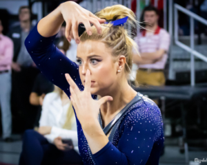A close-up of Gracie Kramer's face. She holds both hands in a contorted pose in front of her face on floor.