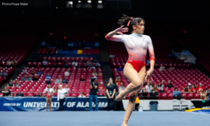 Rutgers gymnast Ella Aird performs a floor routine, mid-dance pose with one arm raised and the other bent gracefully. She wears a red and white ombre leotard in a large arena with rows of red seats and spectators in the background.