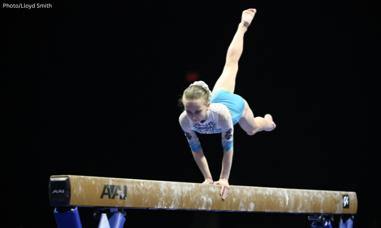 Claire Pease of WOGA performs a handstand on the balance beam during competition, wearing a white and light blue leotard with black and silver detailing against a dark background.
