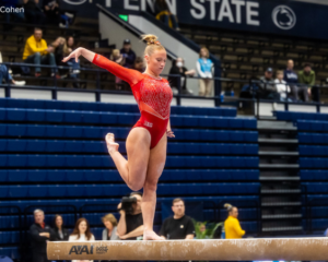A female gymnast in a shimmering red leotard, with her hair in a bun, performs on a balance beam. She is barefoot, balanced on one leg with the other leg bent and raised behind her, and her arms extended to her sides for balance. The background shows a blurry audience in a large arena with "PENN STATE" visible on the wall.