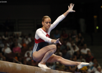 ymnast Terin Humphrey performs a low balance move on the beam, extending one leg forward and both arms for control. She wears a red, white, and navy blue leotard with white beam shoes, focused intently during her routine in a large arena with a blurred audience in the background.
