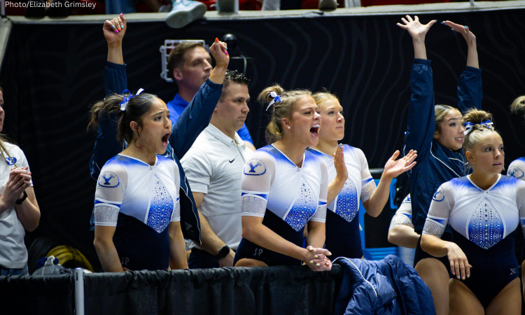 Members of the BYU gymnastics team cheer enthusiastically from the sidelines during a meet. The gymnasts wear white and blue leotards with rhinestone designs, with some raising their arms and others shouting in support.