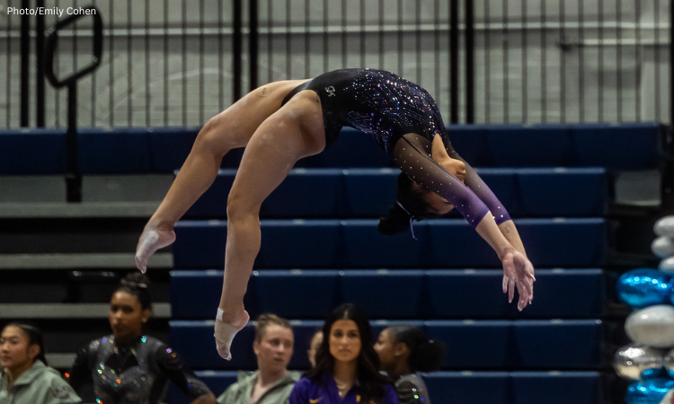 An LSU gymnast performs a back handspring on beam during the NCAA regionals finals. She wears a black leotard with purple and silver sparkles, and blurred teammates and coaches are visible in the background.