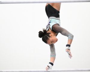 Hezly Rivera performs a handstand on the uneven bars, gripping the lower bar with one hand. She wears a black, mint green, and gray sparkly leotard, with her hair styled in braids pulled into a bun.
