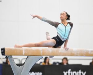 Hezly Rivera performs a balance beam routine, holding a graceful pose with one leg extended and arms poised. She wears a light blue and black leotard with rhinestone detailing. Spectators watch from the background. Photo by Lloyd Smith.
