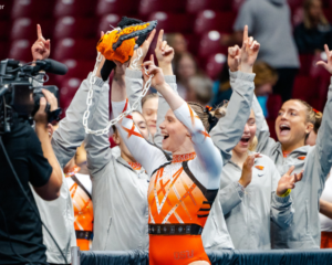 Oregon State gymnast Jade Carey smiles while holding the team’s signature chainsaw spirit stick, also called the Dam Stick. She wears an orange and black leotard, with a blurred arena crowd in the background.