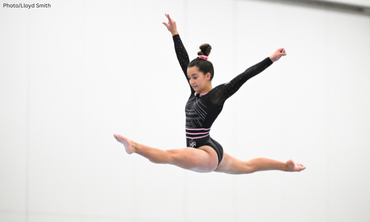 Gymnast Gabby van Frayen performs a split leap in midair during a floor routine, arms extended and toes pointed, wearing a black leotard with pink and silver accents.