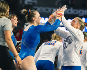 Boise State gymnasts celebrate after a routine, smiling and high-fiving each other while wearing blue leotards and team warm-ups during a meet.