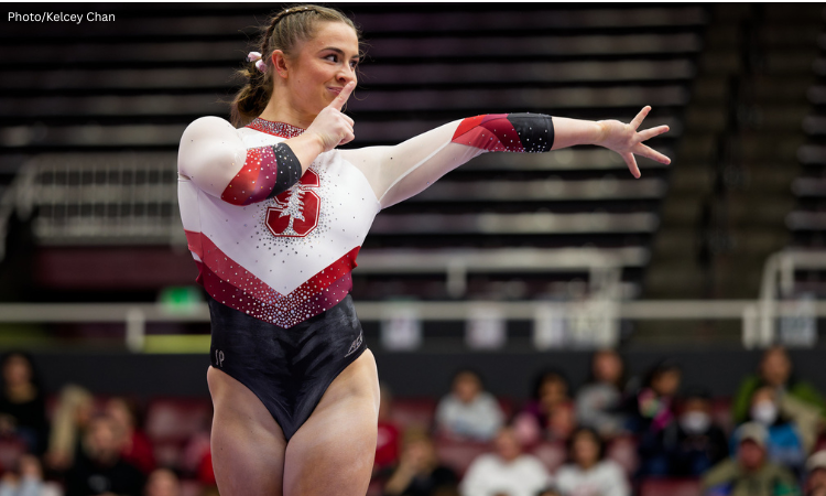 Anna Roberts, a female gymnast, is wearing a red, white, and black long-sleeved leotard with the Stanford University logo on the front. She is standing with her left arm extended to her side and her right hand near her face, with one finger over her lips in a "shhh" gesture. She is smiling and looking off to the side. The background shows a blurred audience in a stadium.
