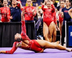 A gymnast poses on floor laying on her left side with her right arm up in the air and her head tilted back. She wears a red leotard and has a red and white ribbon in her blonde hair which is tied in a bun. Her teammates cheer from the sidelines.