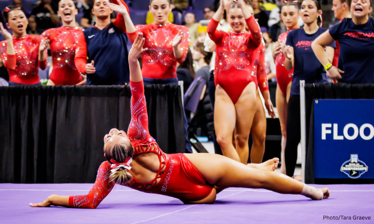 A gymnast poses on floor laying on her left side with her right arm up in the air and her head tilted back. She wears a red leotard and has a red and white ribbon in her blonde hair which is tied in a bun. Her teammates cheer from the sidelines.