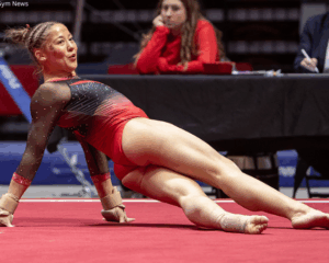 Ava Molina, a female gymnast at Ball State, is wearing a red and black ombré leotard with sheer, sparkling sleeves and wrist guards performing a floor routine on a red mat. She is leaning back on her hands with her legs extended to the side, looking toward the camera with an expressive smile. In the blurred background, a person in a red sweater and a judge at a table watch the performance.