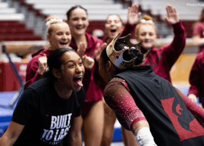A team cheers on in the background as a gymnast celebrates with a teammate. The gymnast wears a crown and a cape, and faces her teammate who stuck out her tongue and smiles.