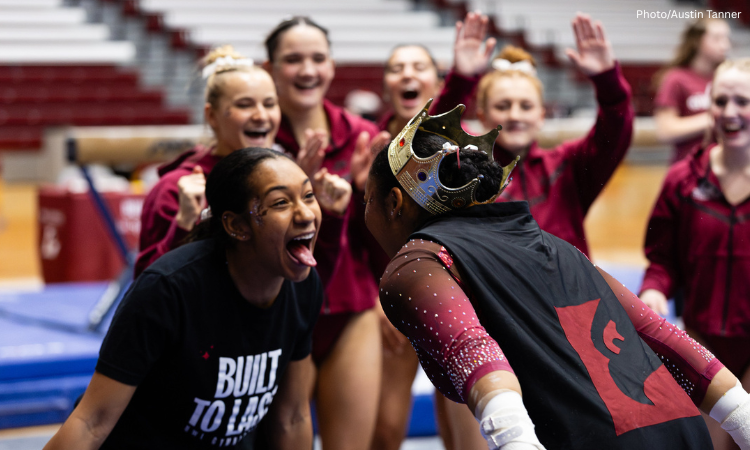 A team cheers on in the background as a gymnast celebrates with a teammate. The gymnast wears a crown and a cape, and faces her teammate who stuck out her tongue and smiles.