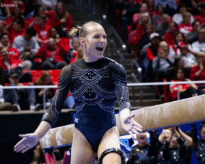 A female gymnast, wearing a long-sleeved black leotard decorated with silver rhinestones, stands smiling with her arms outstretched in front of a balance beam. She has her hair pulled back and is looking up and slightly to her left with an expression of excitement and triumph. In the background, a crowd of spectators is visible in the stands.