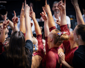 Members of the Oklahoma women’s gymnastics team huddle closely together in a circle, all raising one finger high in the air in unison. The gymnasts wear red sparkly leotards and team hair ribbons, while photographers and staff look on in the background.