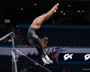 A gymnast flips upside down between the uneven bars against a dark arena background. Her hands reach out in front of her. She wears a black leotard with white accents, her hair is up in a bun.