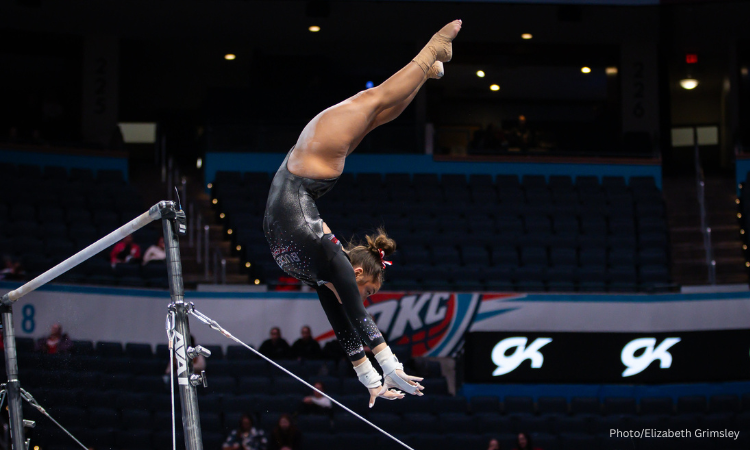 A gymnast flips upside down between the uneven bars against a dark arena background. Her hands reach out in front of her. She wears a black leotard with white accents, her hair is up in a bun.