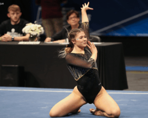 A gymnast poses on her knees on the floor with one hand crossing in front of her and the other up in the air. Her facial expression is confident. Her hair is braided back into a low bun and she wears a black leotard with yellow detailing.