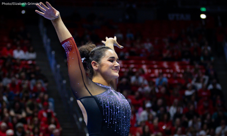 tanford gymnast Sienna Robinson stands on the competition floor with her arms raised in a confident finishing pose, smiling slightly. She wears a black leotard with sheer sleeves and shimmering purple rhinestone designs, while a large arena crowd in red fills the blurred background.