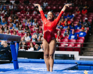 Gabrille Dildy, a gymnast from Rutgers University, wearing a red leotard stands on a blue mat in a large arena, with her arms raised in a "V" shape and a wide smile. She is positioned in front of a balance beam, with a crowd of spectators in red and blue seating visible in the blurred background.